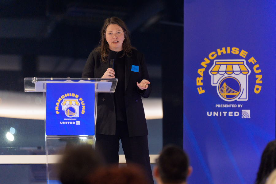 Nina Kaiser speaking at a podium during a Franchise Fund event presented by United. There are two large blue banners with the event logo, which features a storefront and a bridge, and the words "Franchise Fund" in yellow.