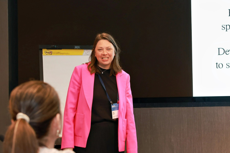 Nina Kaiser in a pink blazer is smiling during a presentation in a conference room. There is a large screen displaying text to her right.
