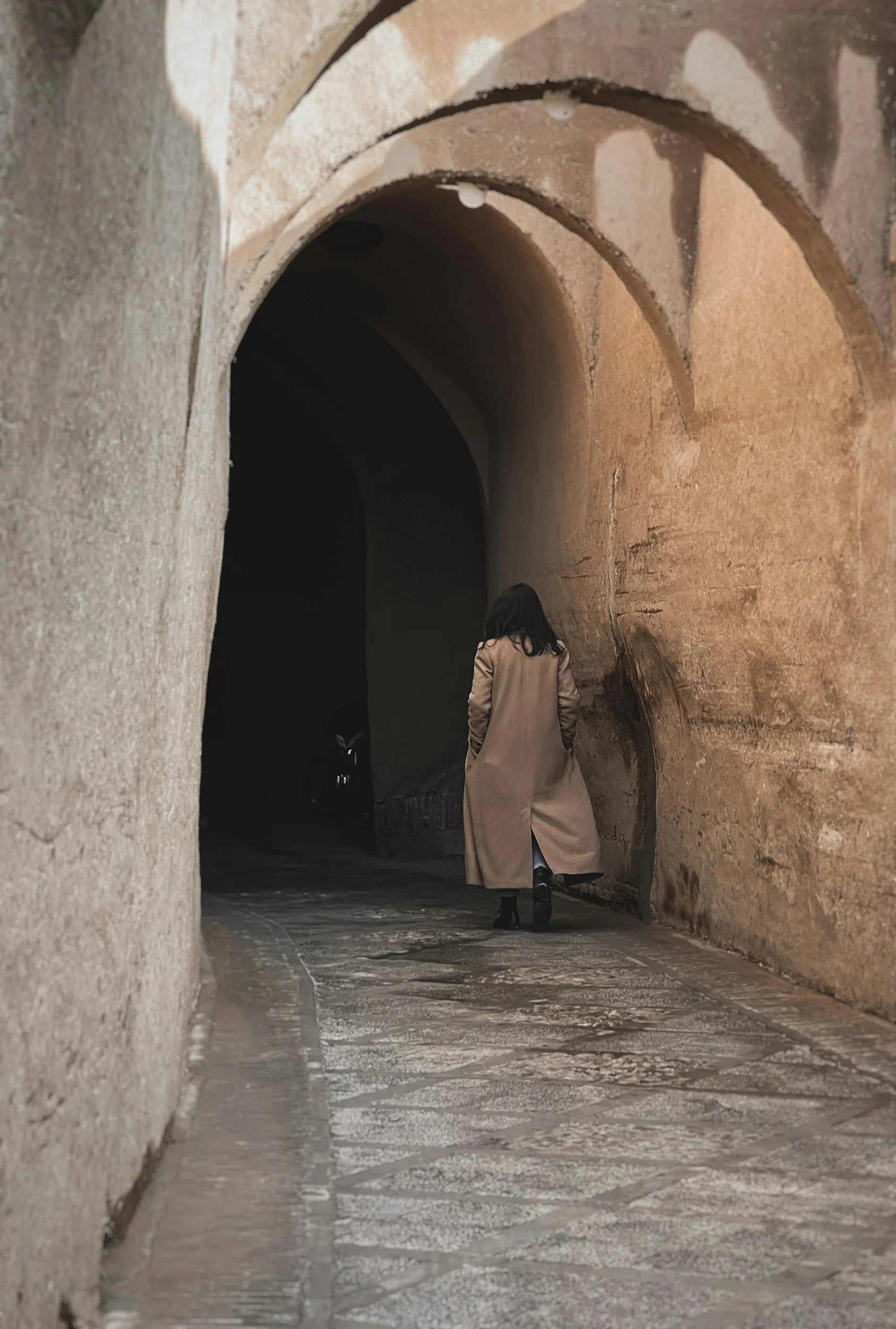 Person in beige coat walking through a stone tunnel with arched ceiling, stone walls, and paved floor.
