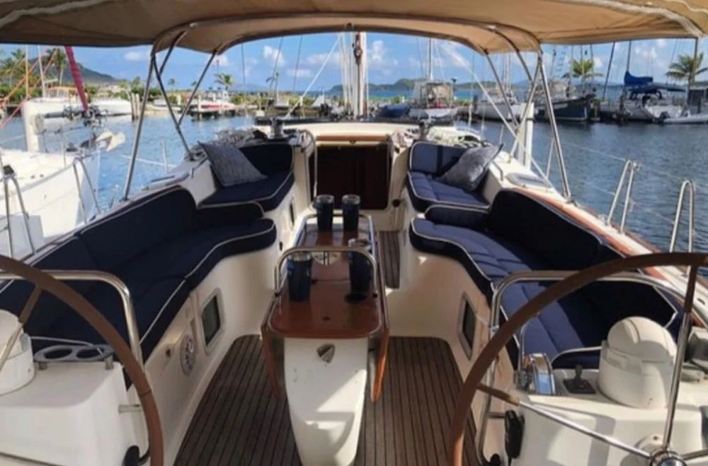 View of a yacht deck with navy blue cushioned seating, a wooden table, and a canopy, docked at a marina with other boats and sailboats in the background.