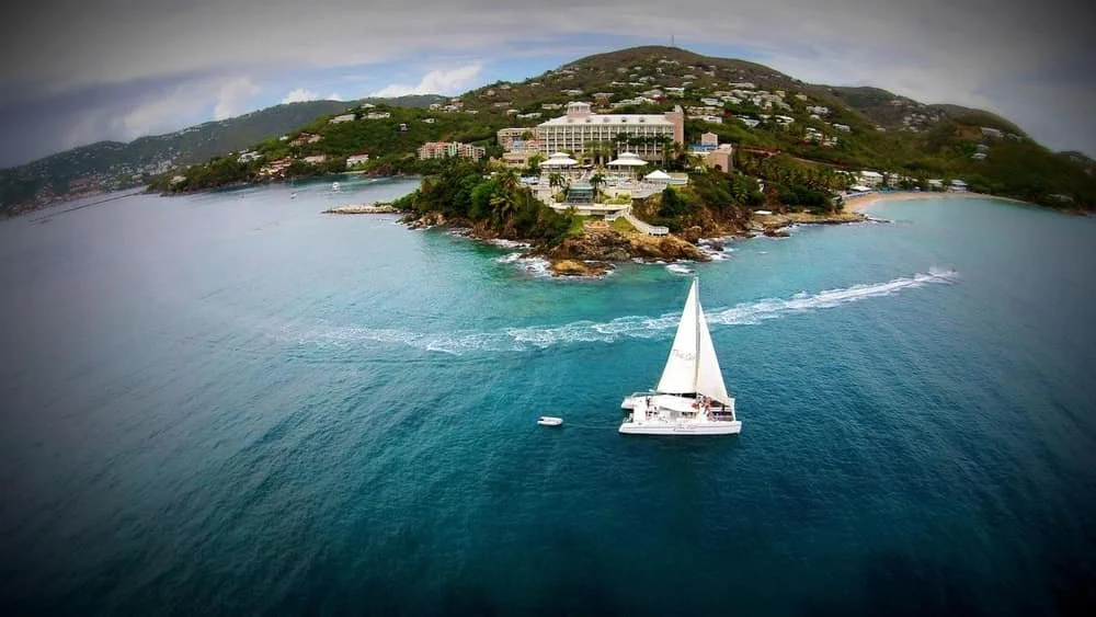 A sailboat cruising in turquoise waters near a lush, green island with large buildings and a hilly landscape.