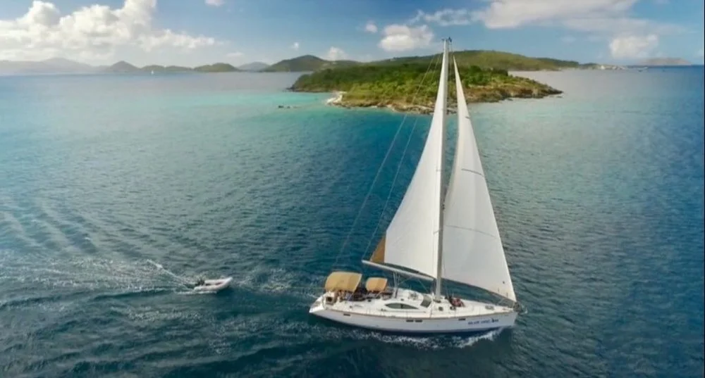 A sailboat with white sails sailing in blue waters near a green island with hills and a cloudy sky.
