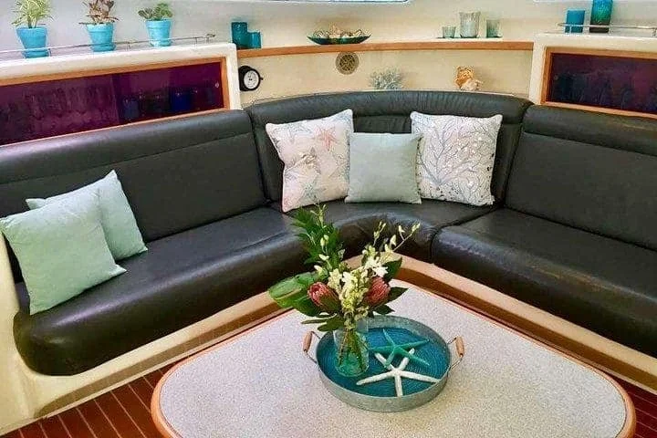 A corner sectional black leather sofa with various pastel pillows, a round coffee table with a flower arrangement and starfish decor, and a shelf with decorative items and teal planters in the background.