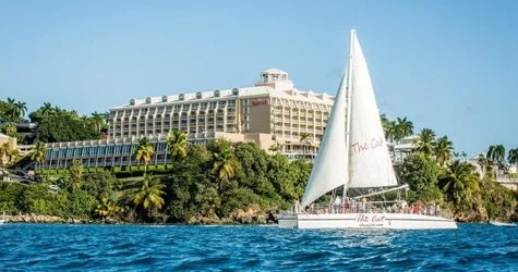 A sailboat on the water near a hilly area with lush greenery and a large hotel or resort building in the background.