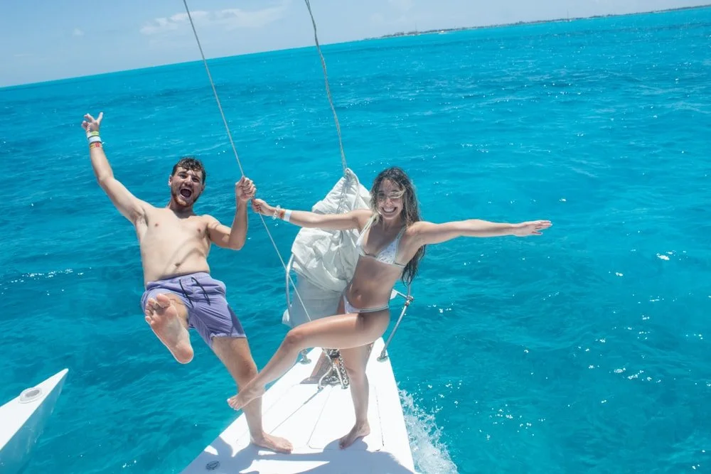 A man and a woman are on a sailboat in clear blue ocean water. They are both smiling and appear to be having fun, with the man jumping and the woman standing on the edge of the boat with her arms outstretched.