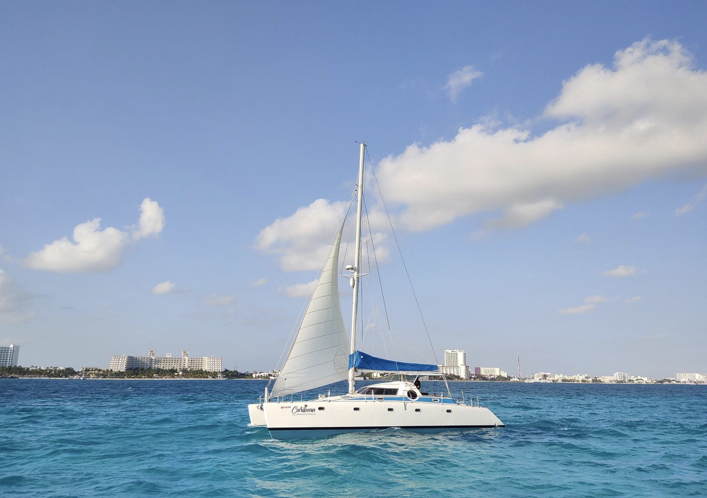 A white sailboat with blue accents floating on turquoise water near a city skyline with various white high-rise buildings and a partly cloudy sky.