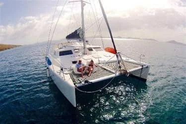 A white catamaran sailing boat with two people relaxing on the deck, surrounded by calm waters under a partially cloudy sky.