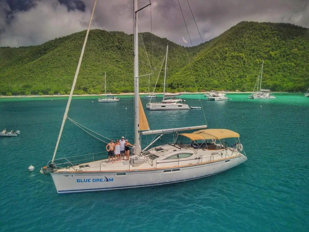 People standing on a sailboat named 'Blue Dream' in a turquoise bay with green mountains in the background. Several other boats are anchored nearby.