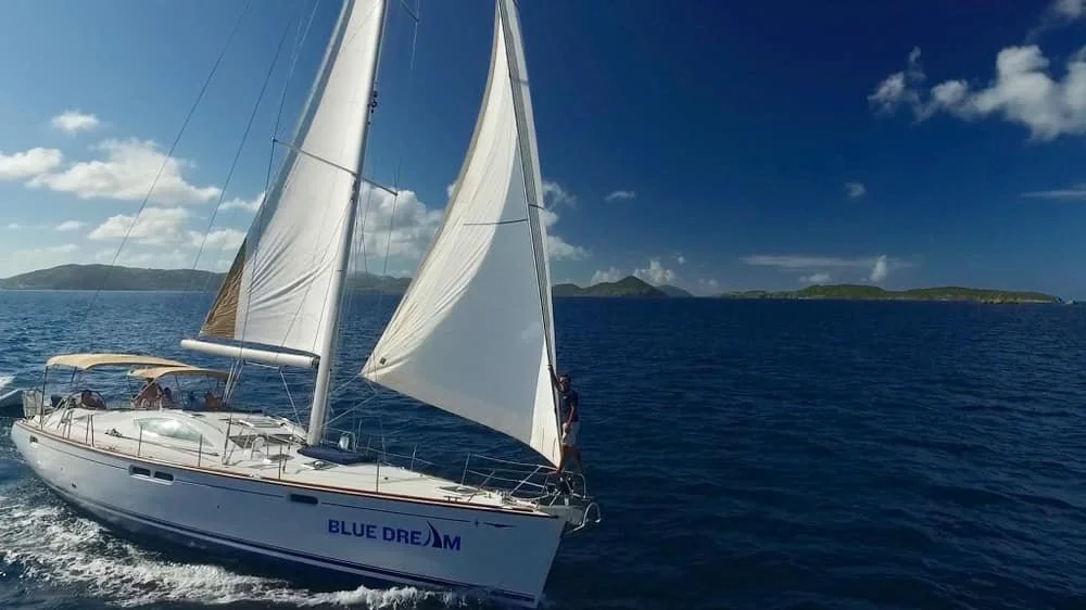 A sailboat named 'Blue Dream' cruising on the ocean with its sails up, while a person stands at the bow. There are some islands in the distance under a partly cloudy sky.