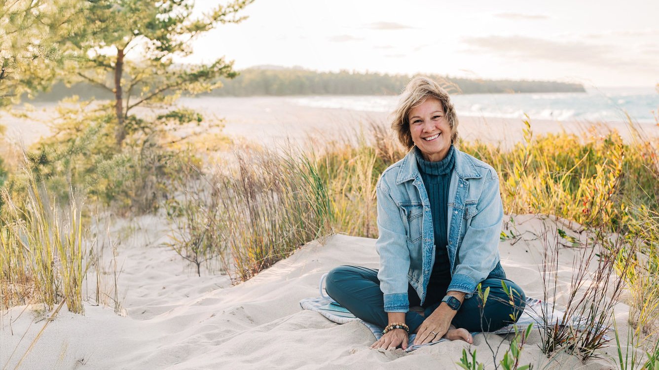 Midlife woman keynote speaker Cathy Fitzgerald sitting on the beach at Lake Michigan