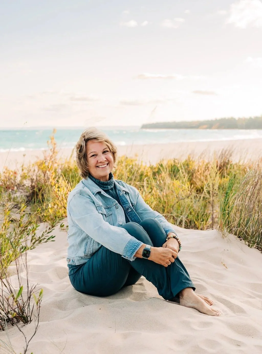 A woman sitting cross-legged on a sandy beach near some grass, smiling at the camera with the ocean and cloudy sky in the background during daytime.