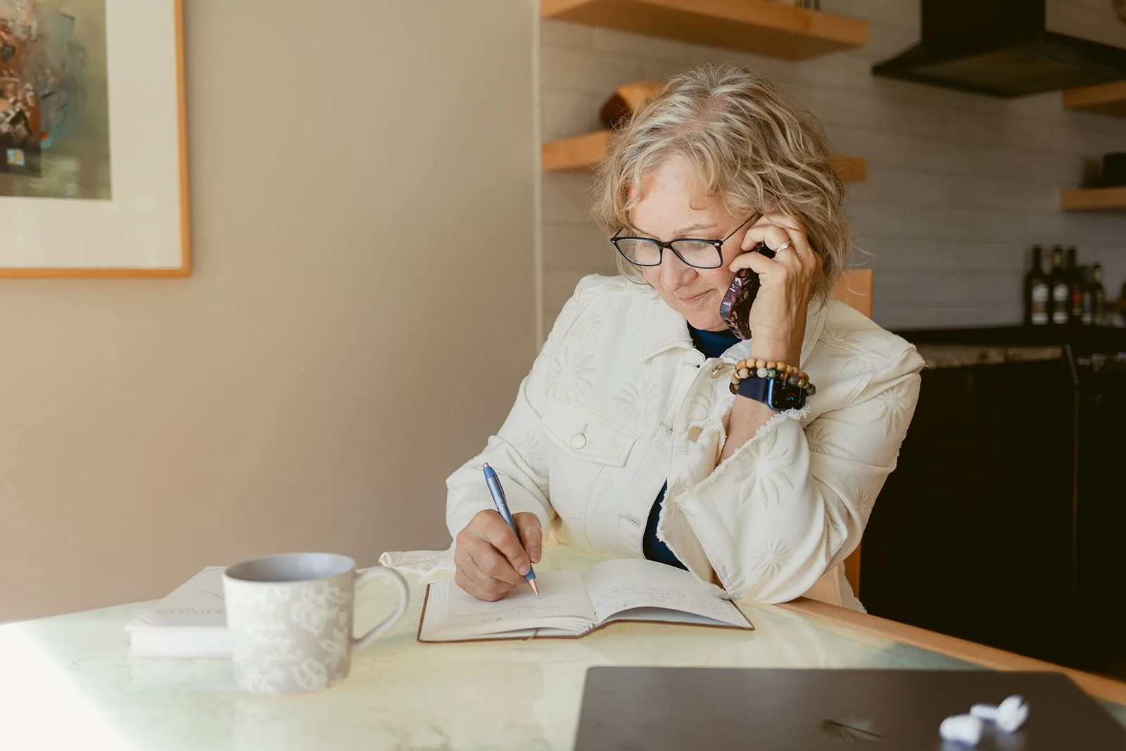 Studio Business coach Cathy Fitzgerald talking on phone and writing in notebook at table.