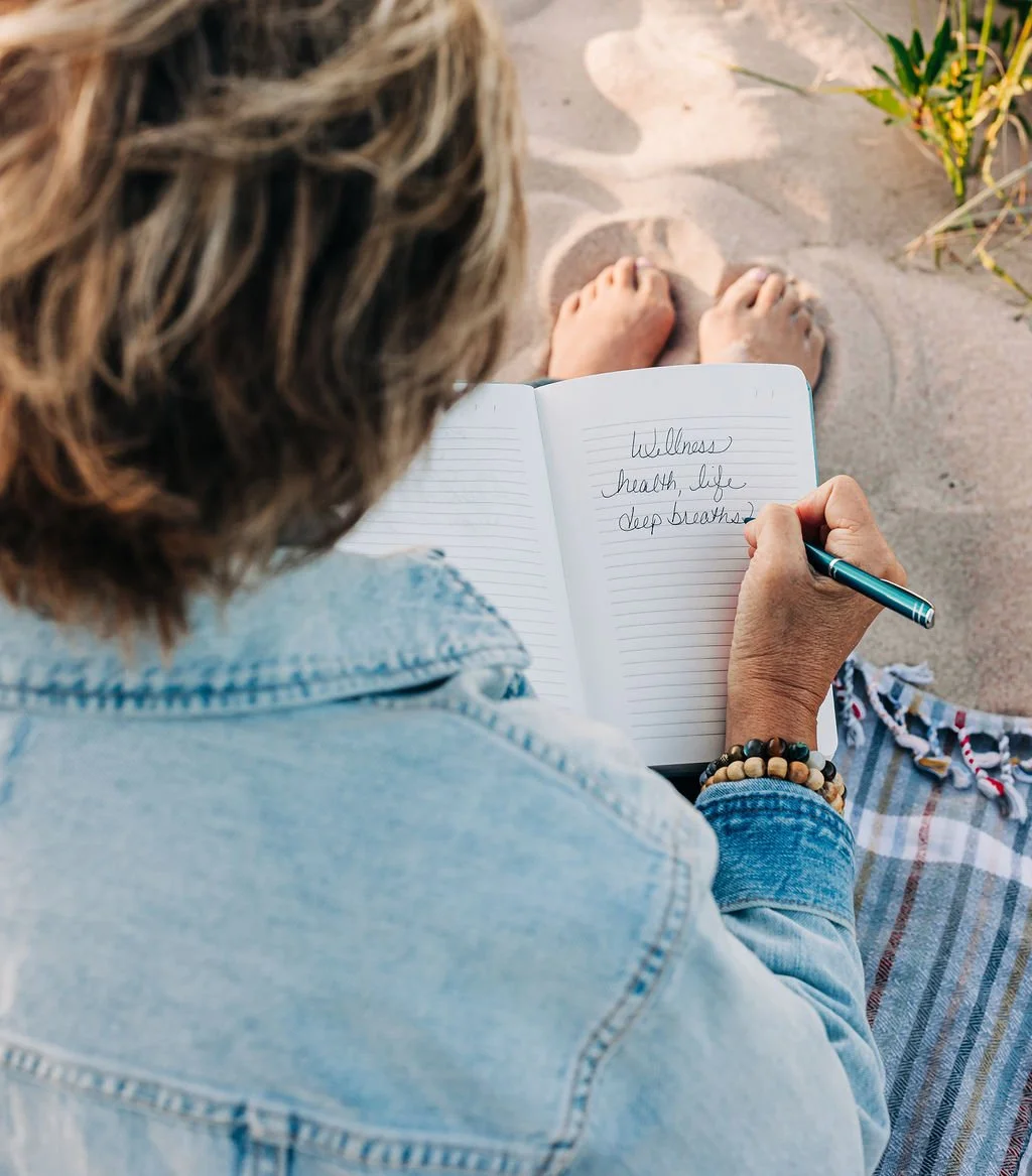 A person sitting on the sand at the beach writing in a notebook with the words "Wellness, health, life, deep breaths" visible on the page.