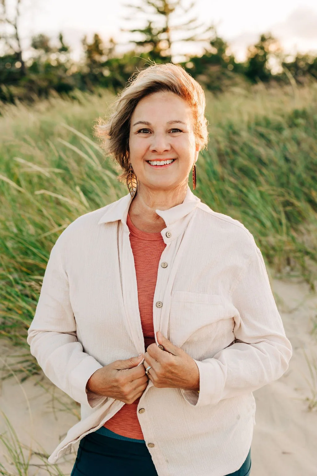 A woman with short hair smiling outdoors in a natural setting with green grass and trees in the background, during golden hour lighting.
