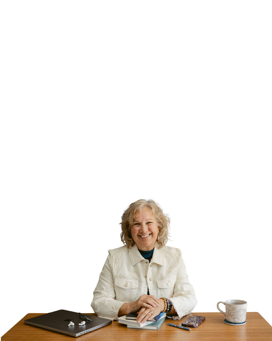 Smiling elderly woman sitting at a wooden desk with a laptop, books, pen, smartphone, and mug. She has curly blonde hair and is wearing a white jacket.
