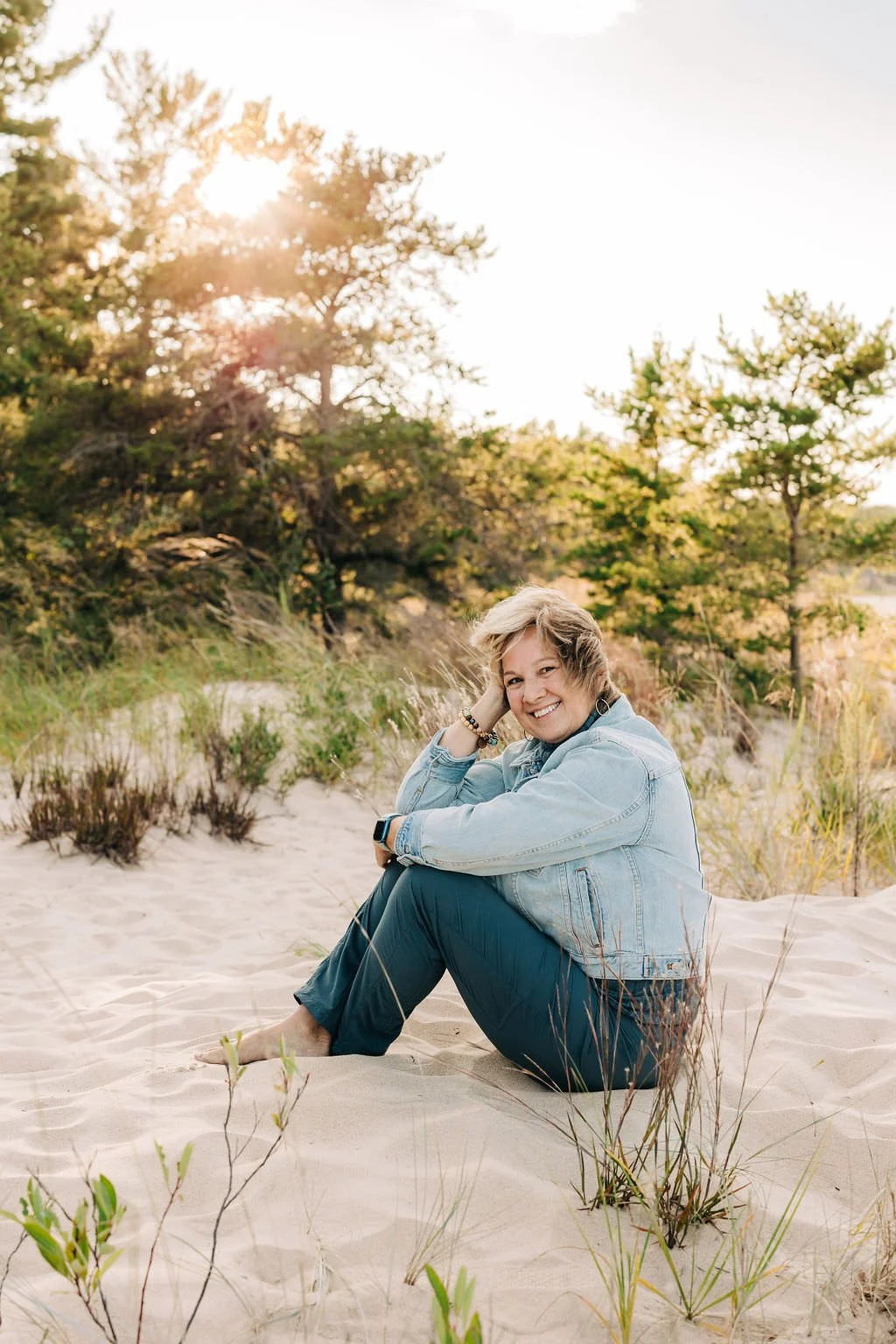 Woman sitting on sandy beach with smiling face, wearing a denim jacket and dark pants, surrounded by sand, grass, and trees, with the sun setting or rising in the background.
