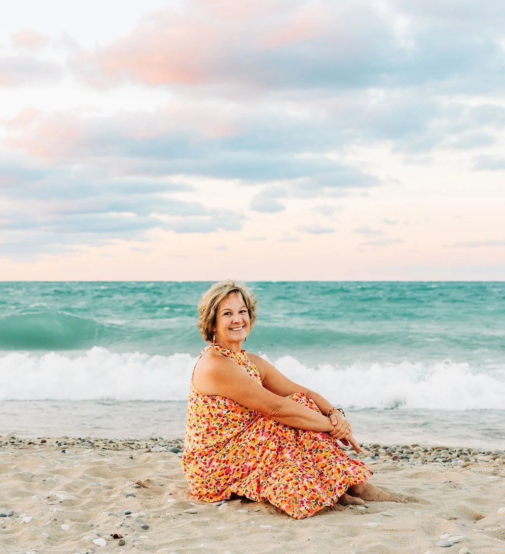 A woman sitting on the sandy beach with the ocean and a cloudy sky in the background, smiling and wearing a colorful floral dress.