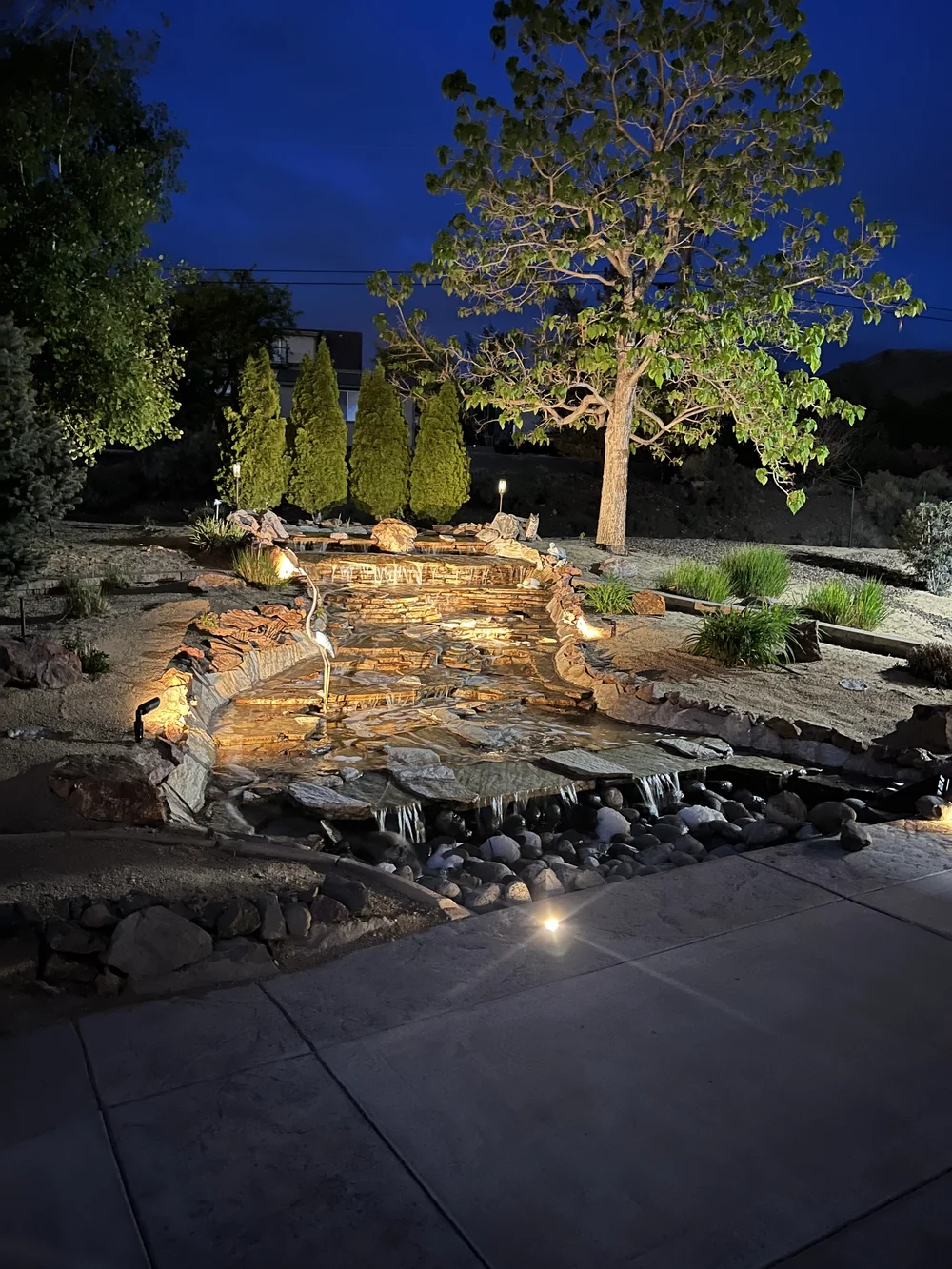 Natural rock waterfall lit at night next to a concrete patio, built by Jefe's Design and Build