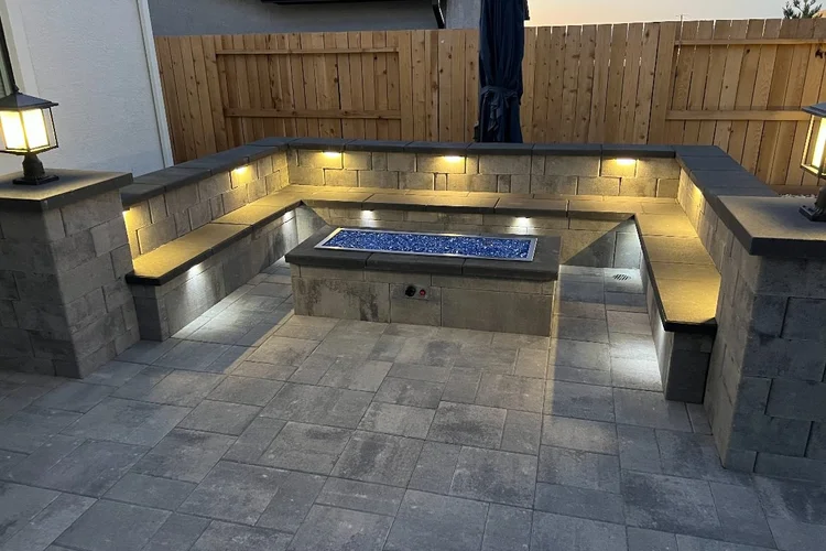 Linear gas fire pit with blue fire glass and a lit seating wall on a paver patio