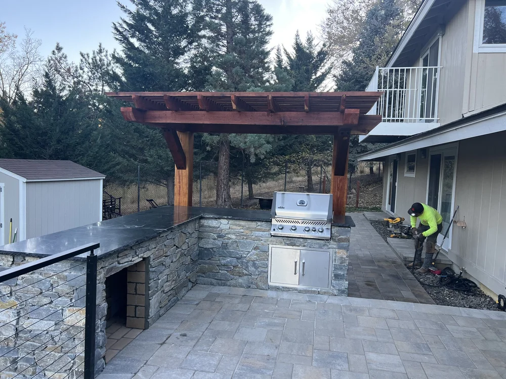 Outdoor kitchen with a stone grill island and a stainless steel grill under a wood pergola, built by Jefe's Design and Build