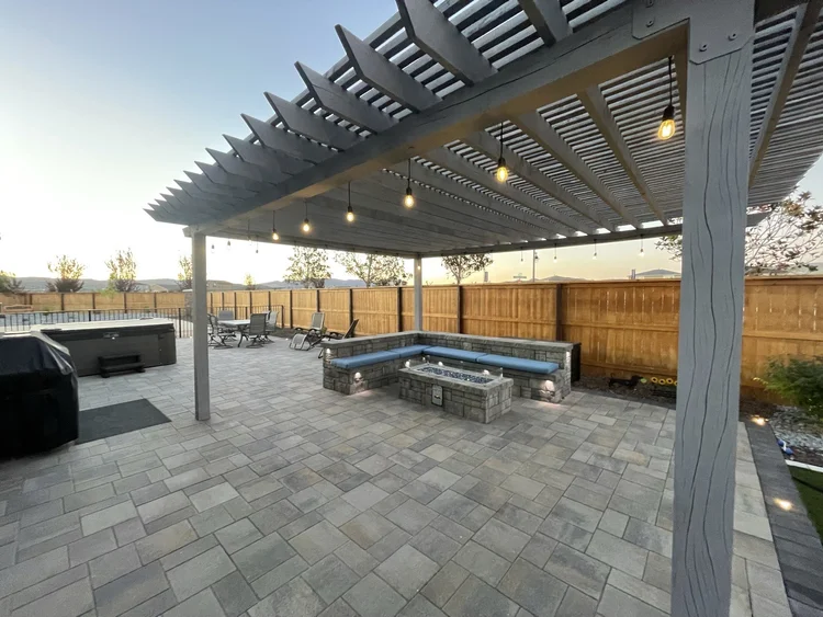 Gray wood pergola with string lights over a paver patio with sectional seating and a fire pit