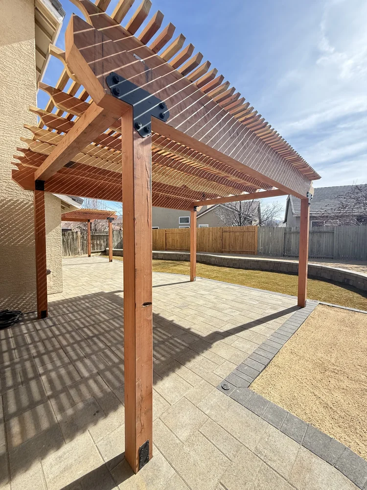Wood pergola with a slat roof over a paver patio next to a low retaining wall