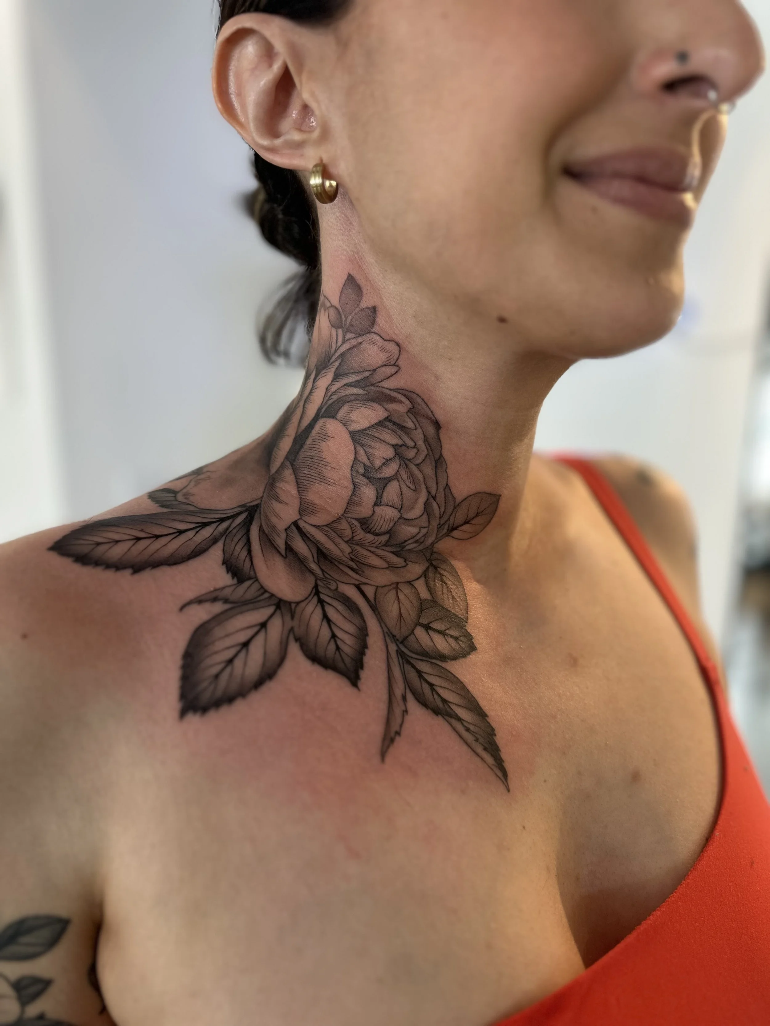 Close-up of a woman with a floral tattoo on her neck and shoulder, wearing a gold earring and a nose ring, smiling and dressed in a red sleeveless top.