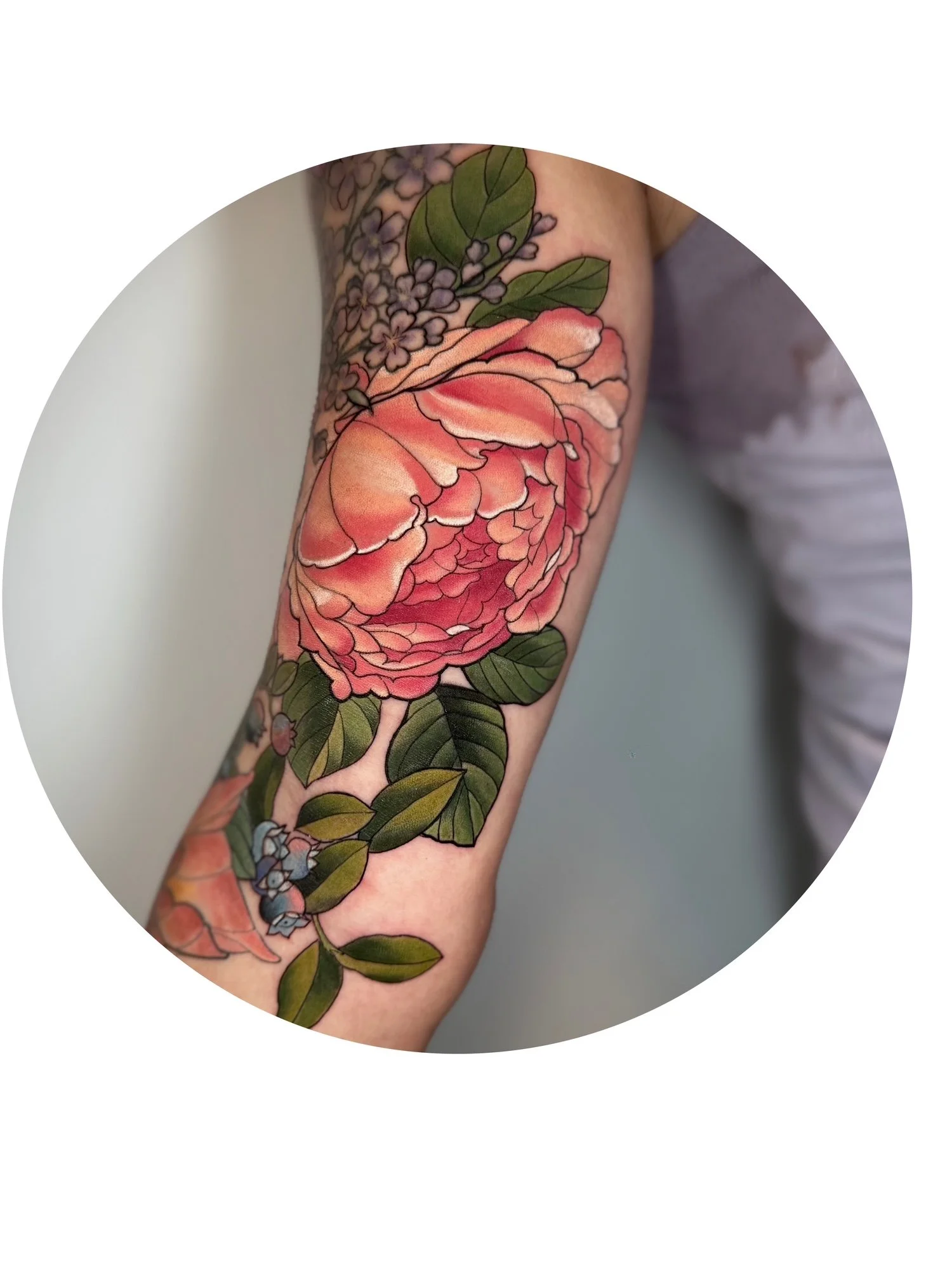 Colorful floral tattoo of a pink peony blossom with green leaves and small purple flowers on a person's arm.