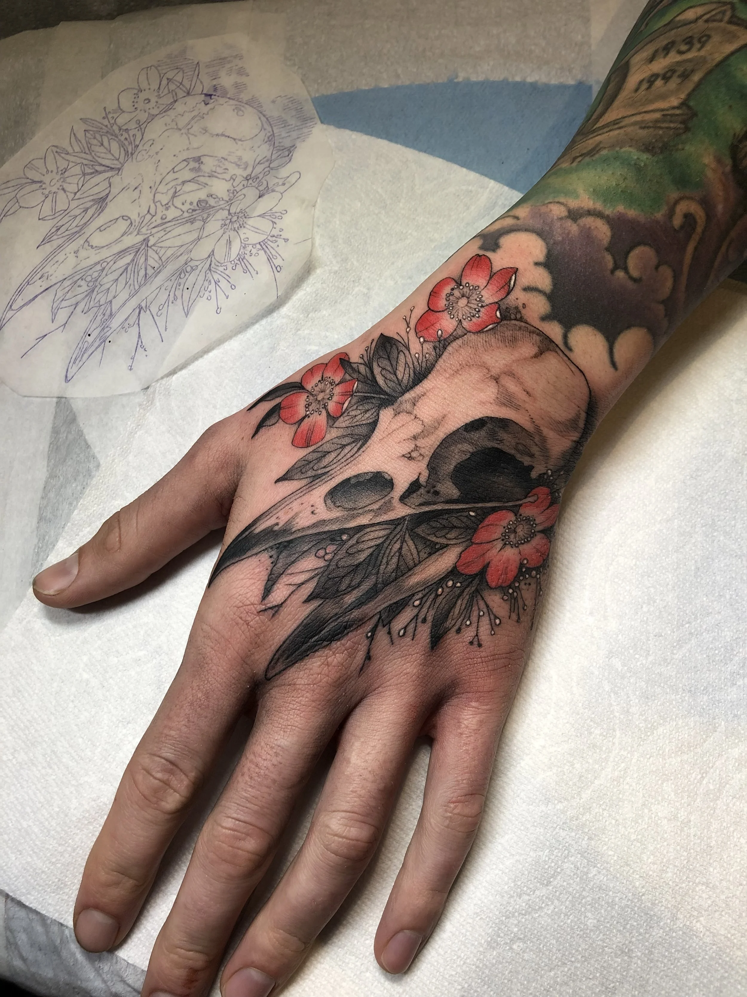 A tattoo of a human skull surrounded by red flowers and black leaves on a person's hand and wrist.