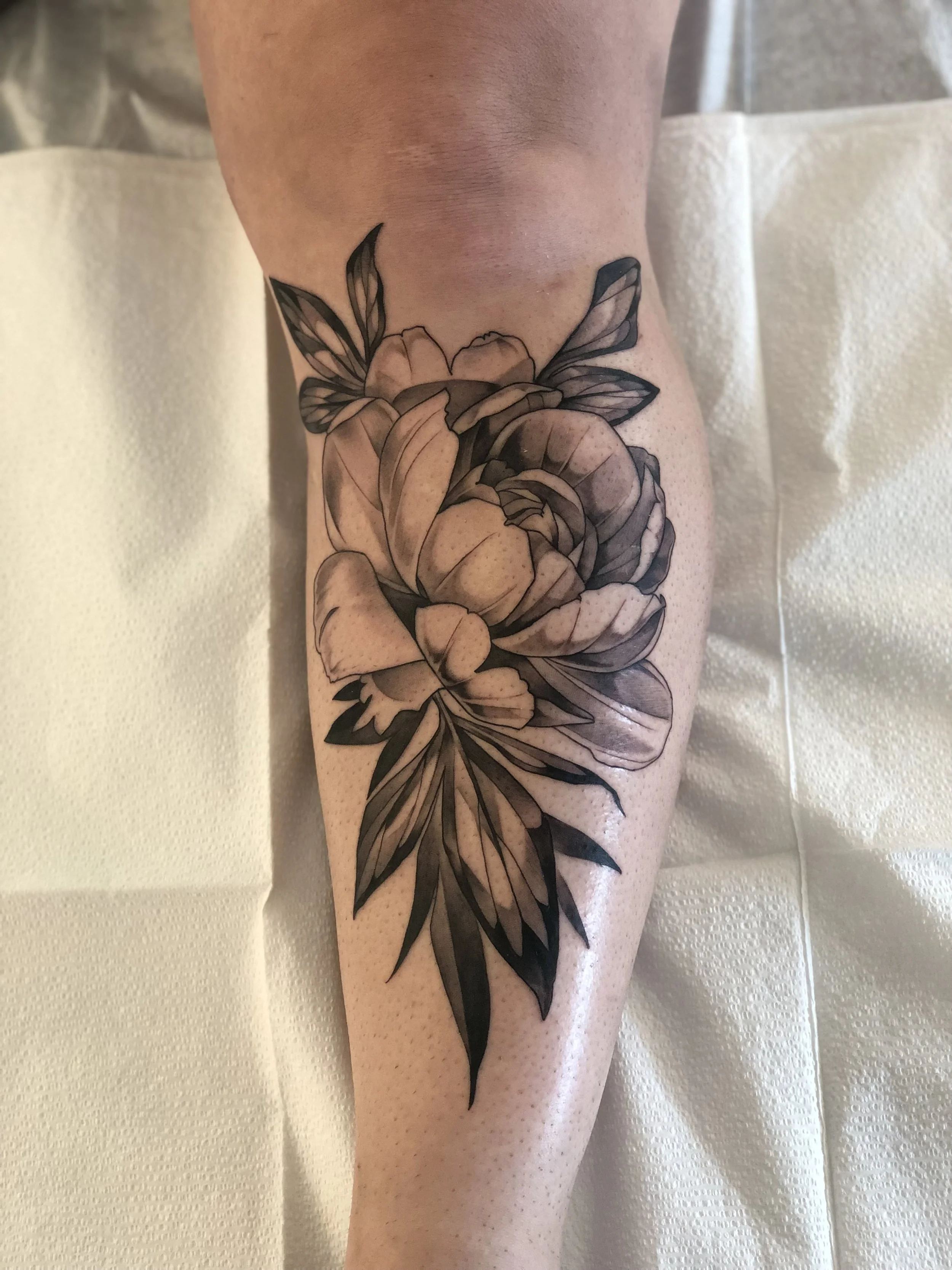 Black and gray floral tattoo on person's calf featuring a large blooming flower, surrounded by leaves and smaller floral details.