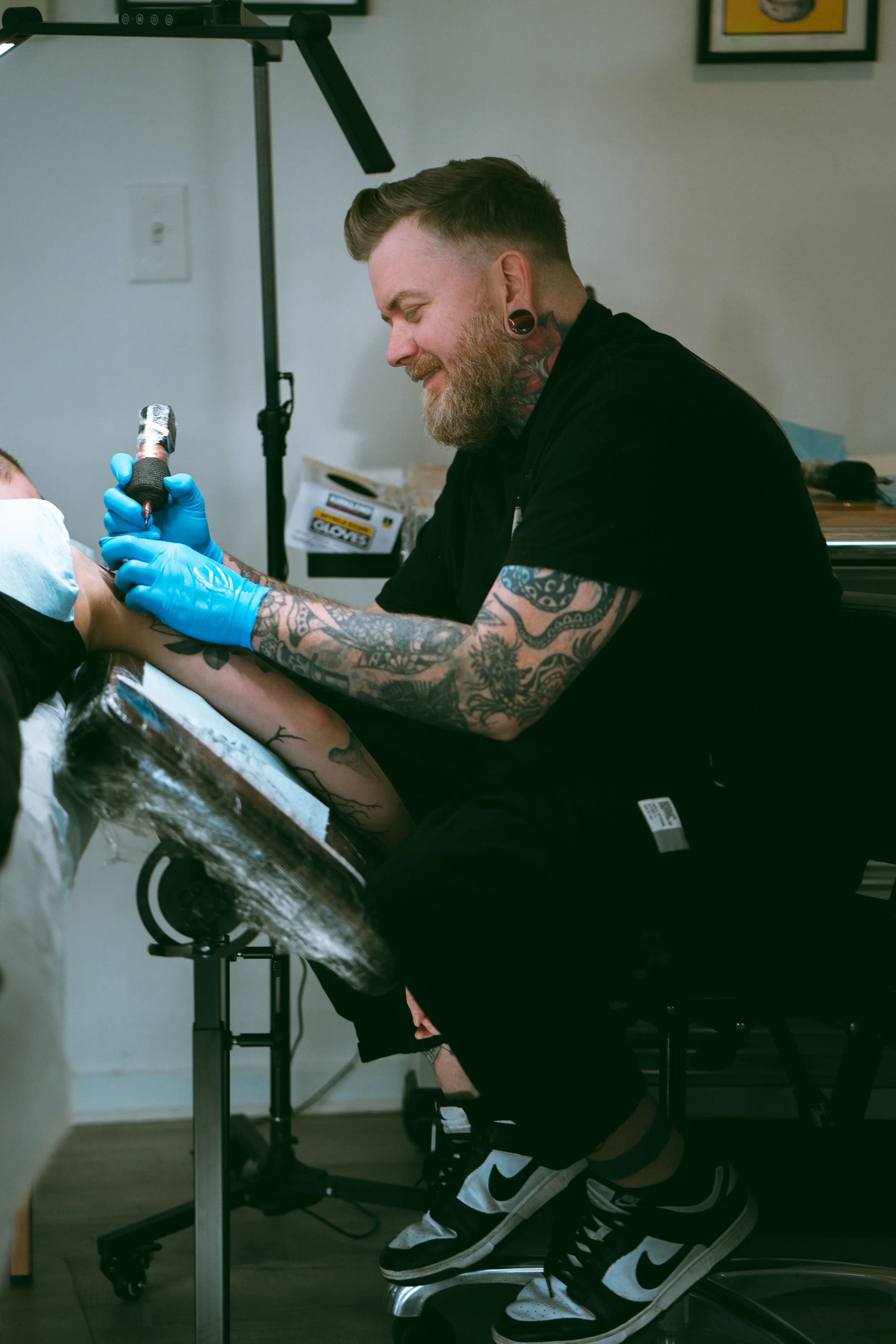 A tattooed man with gloves tattooing a person's arm in a tattoo studio.