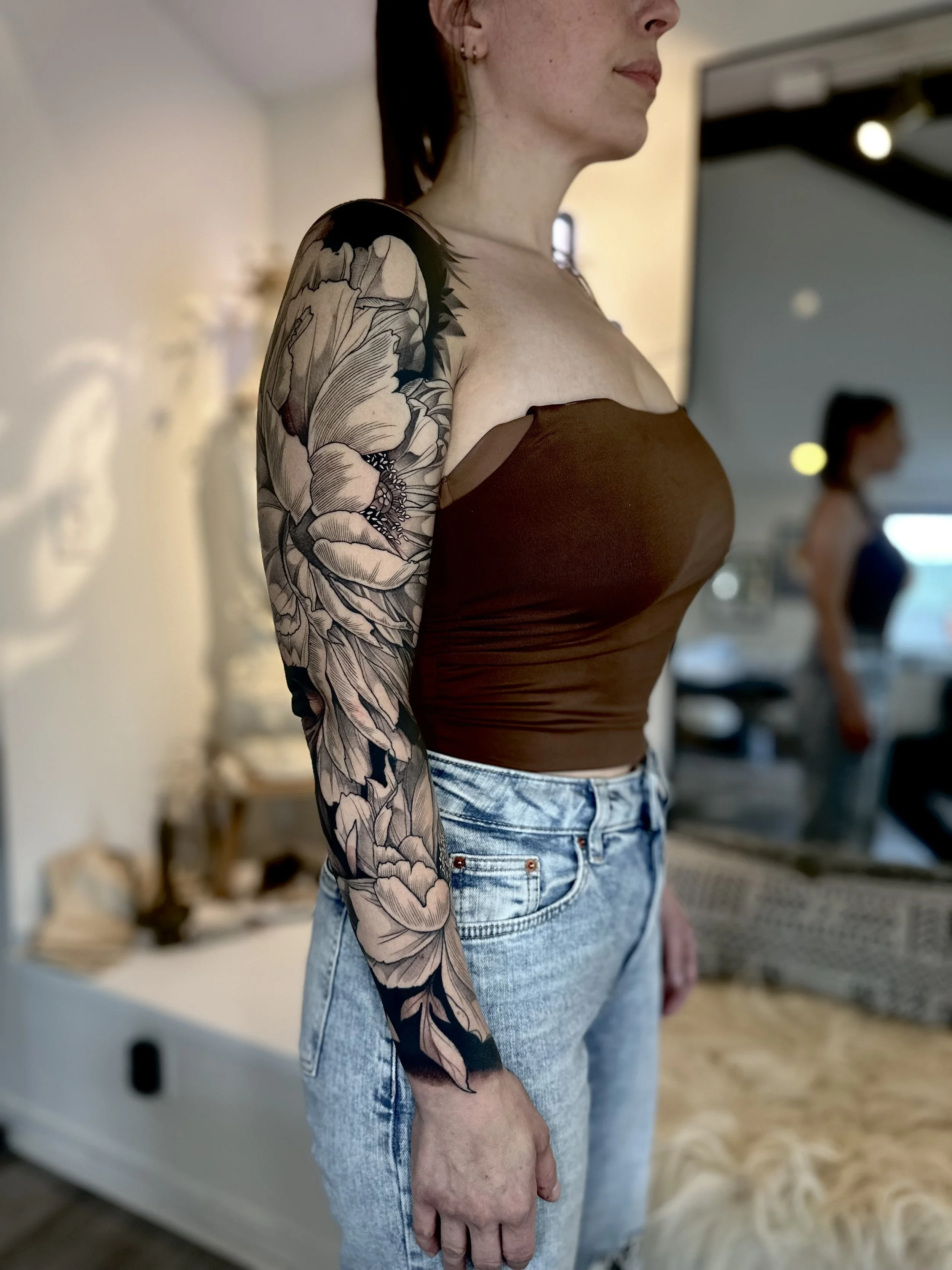 Woman with a floral tattoo sleeve on her left arm, wearing a brown strapless top and high-waisted light blue jeans, standing indoors.