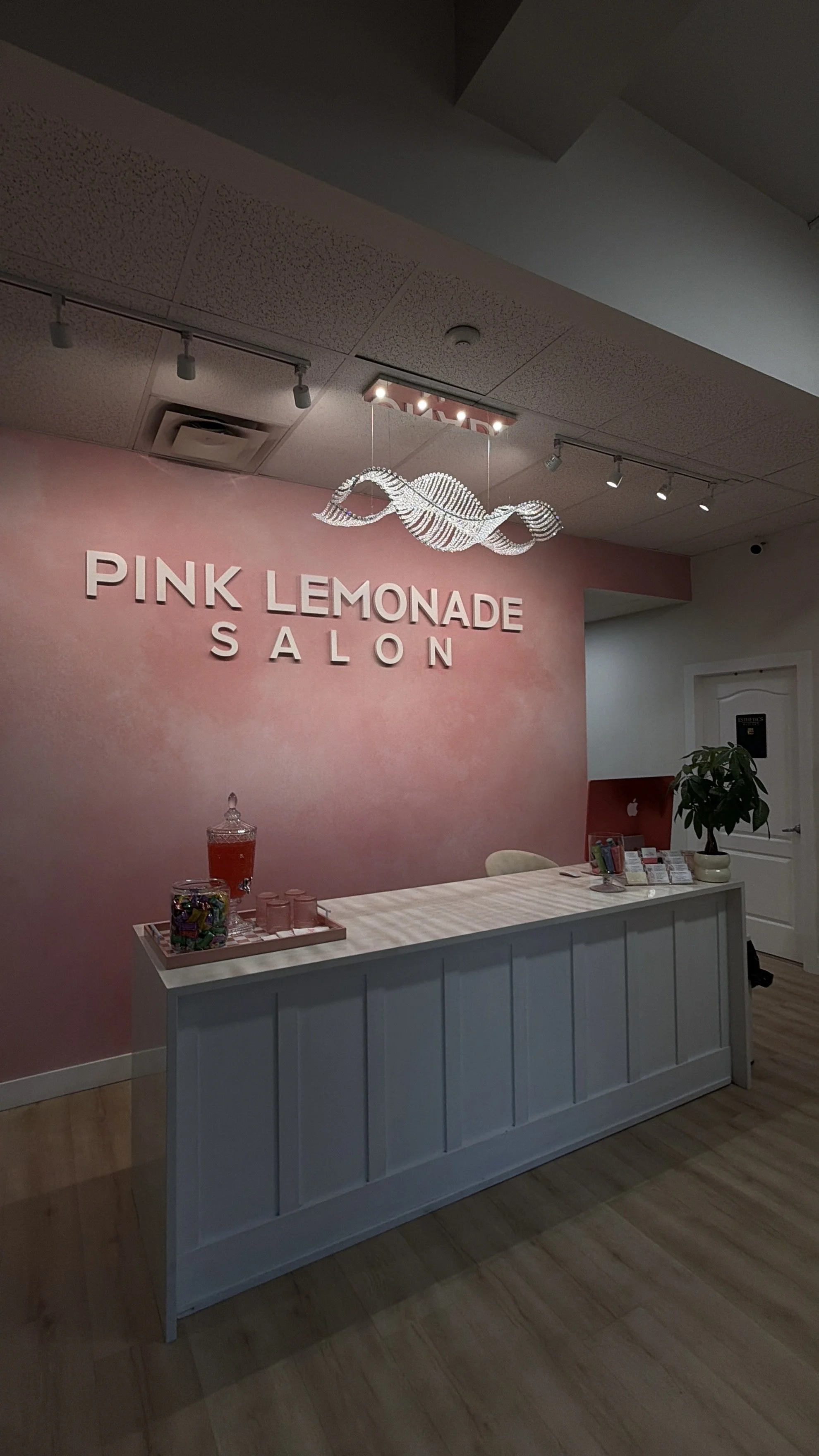 Interior of Pink Lemonade Salon with pink wall, white reception desk, beach-themed chandelier, potted plant, and candy jars.
