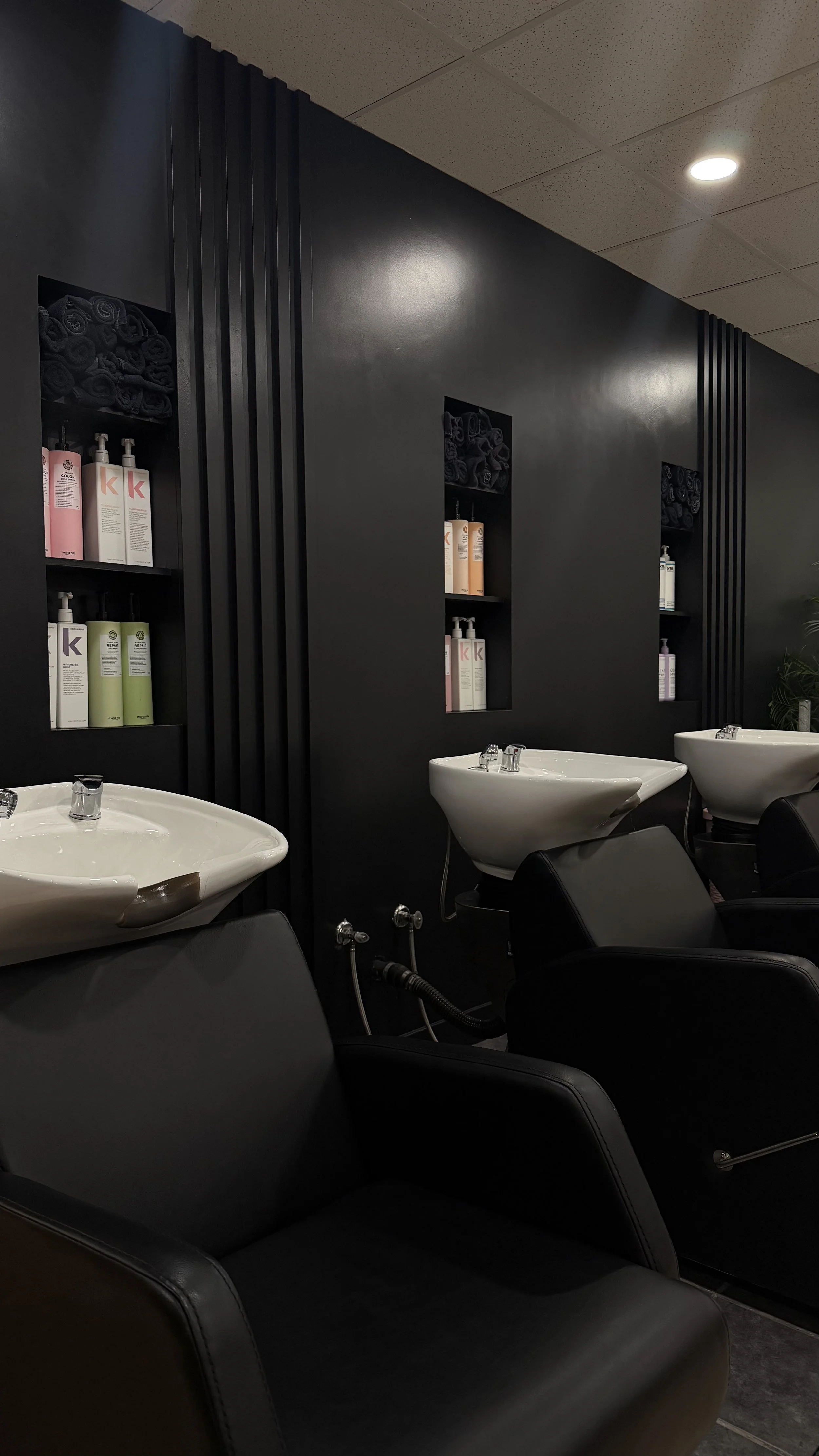 Interior view of a hair salon with black wall, shelves with towels and hair products, and white washbasins with chairs.
