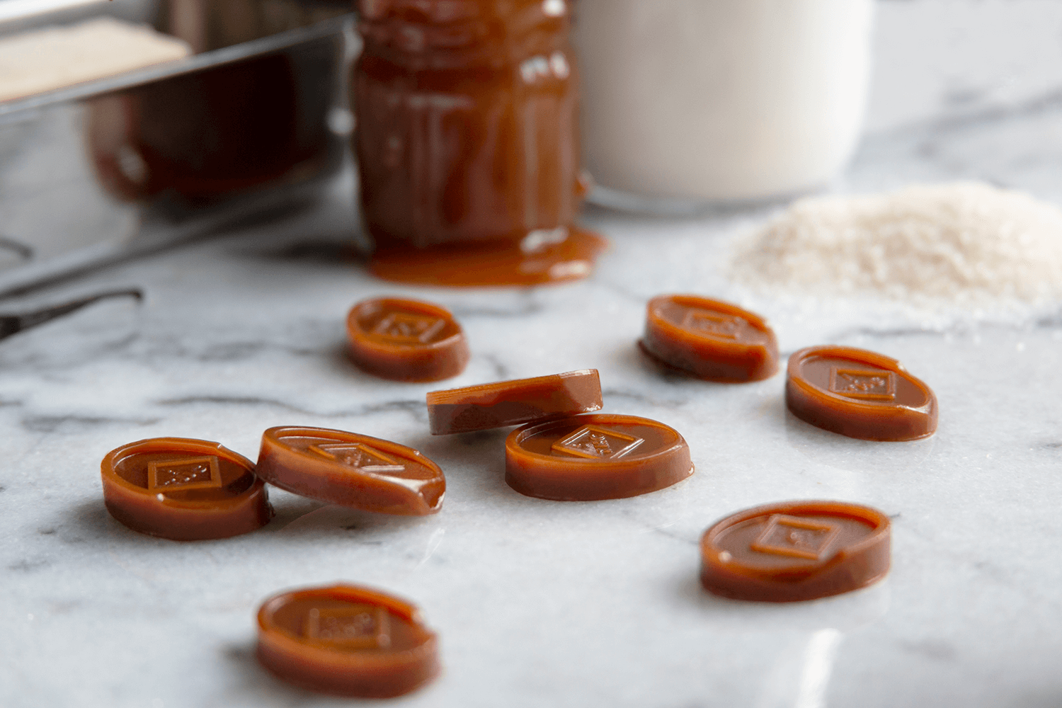 Caramel chocolates on a marble surface with a jar of caramel, a white jar of cream, shredded coconut, and a baking pan in the background.