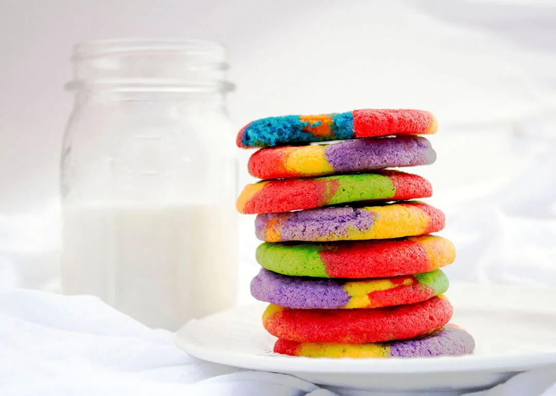Colorful rainbow cookies stacked on a white plate with a glass jar of milk in the background.