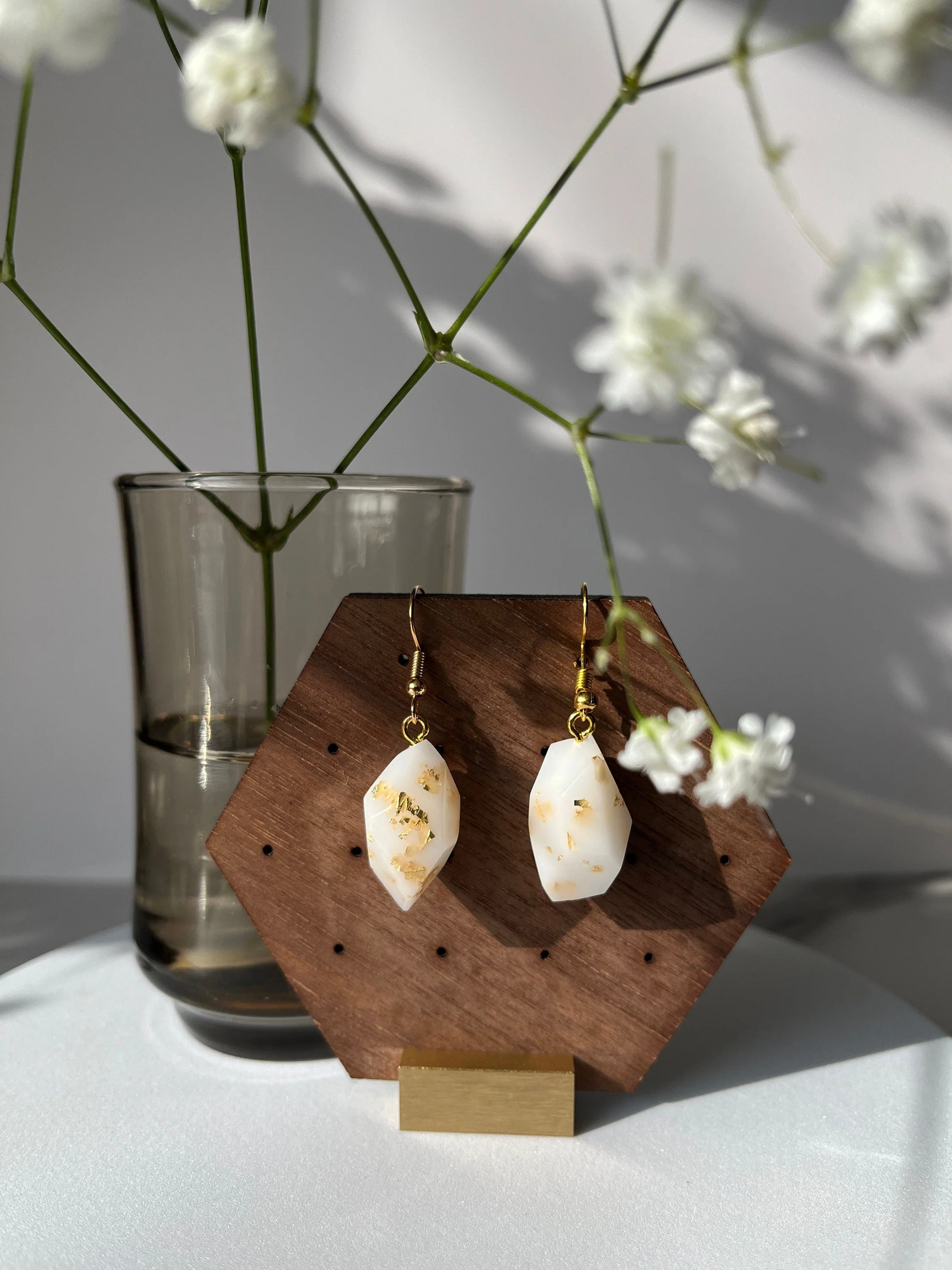 White and gold earrings hanging on a wooden jewelry display, with a clear glass vase holding white flowers in the background.