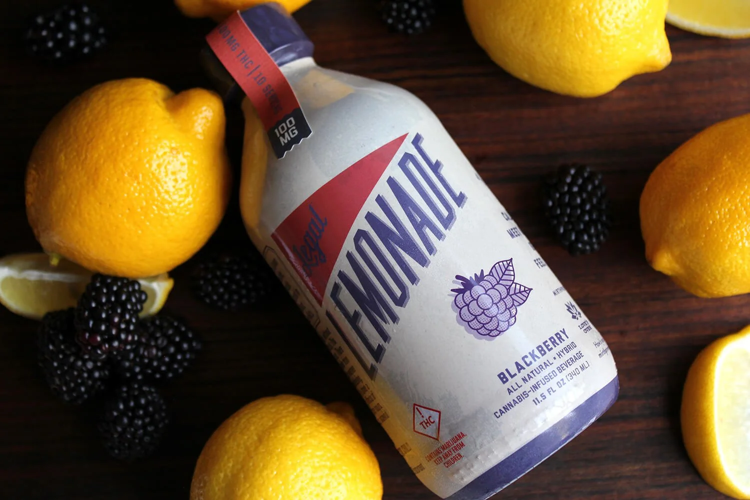 A bottle of Lemonade CBD blackberry flavored surrounded by lemons, lemon slices, blackberries, and a dark wooden surface.