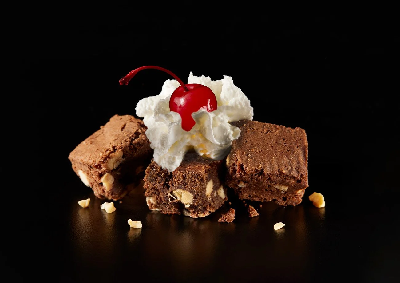 Three brownie squares topped with whipped cream and a cherry on a black background.