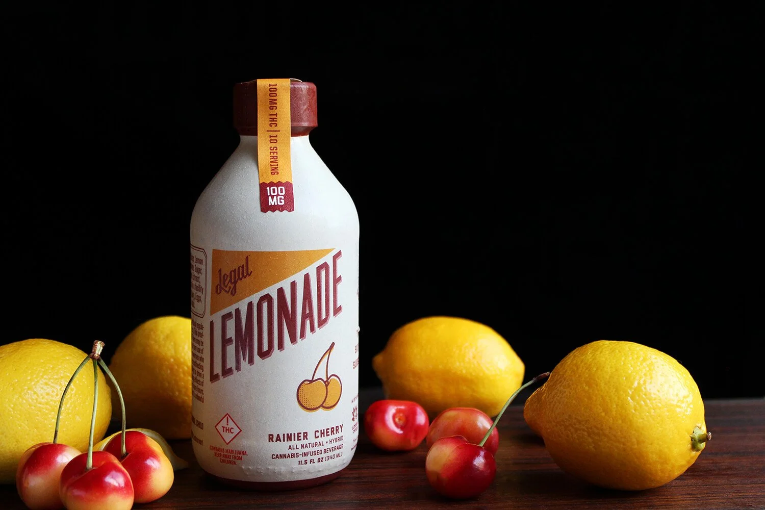 A bottle of legal lemonades flavored with rainier cherry, surrounded by lemons and cherries on a wooden surface against a black background.