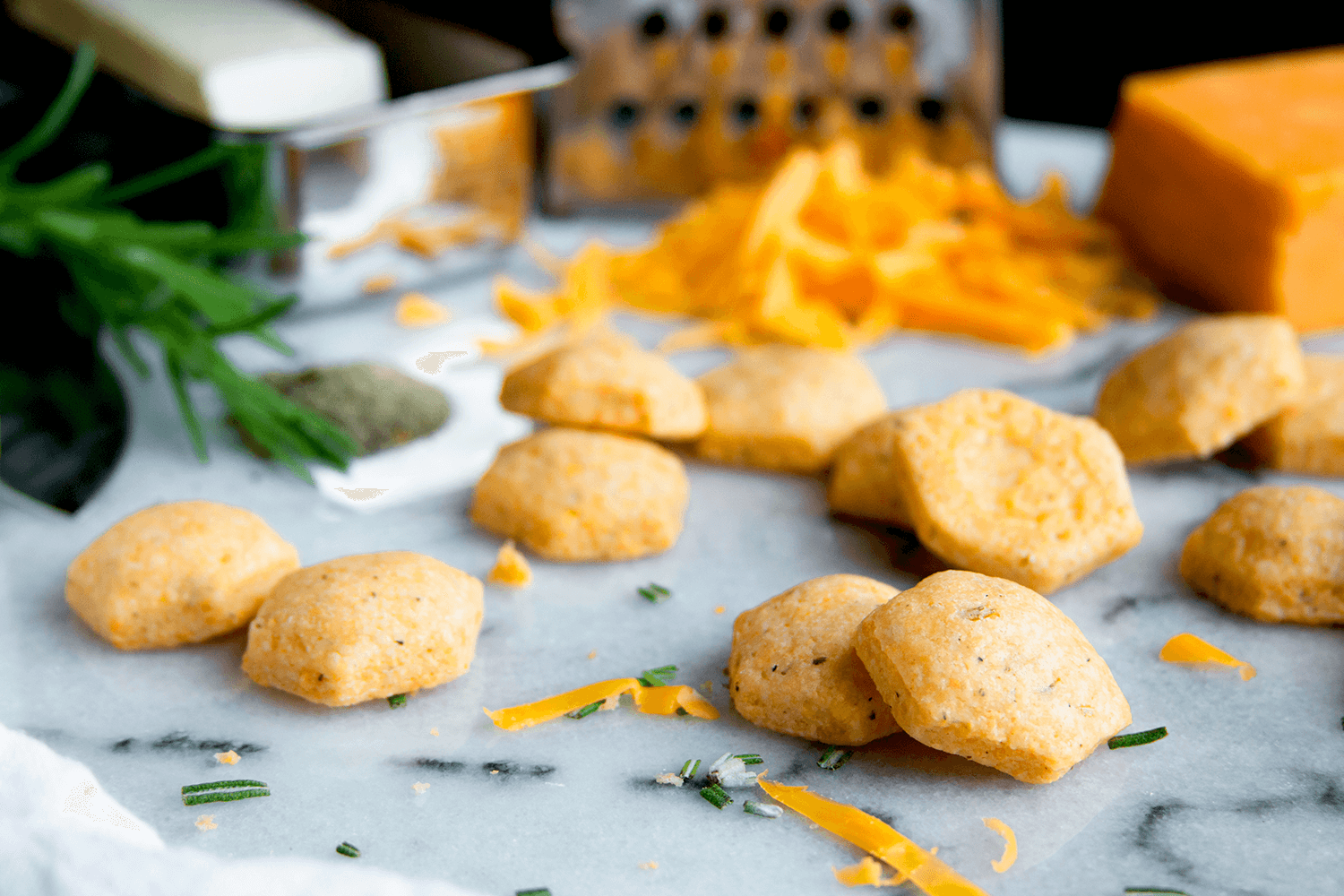 Close-up of homemade cheese curds on parchment paper, with shredded cheese and herbs in the background.