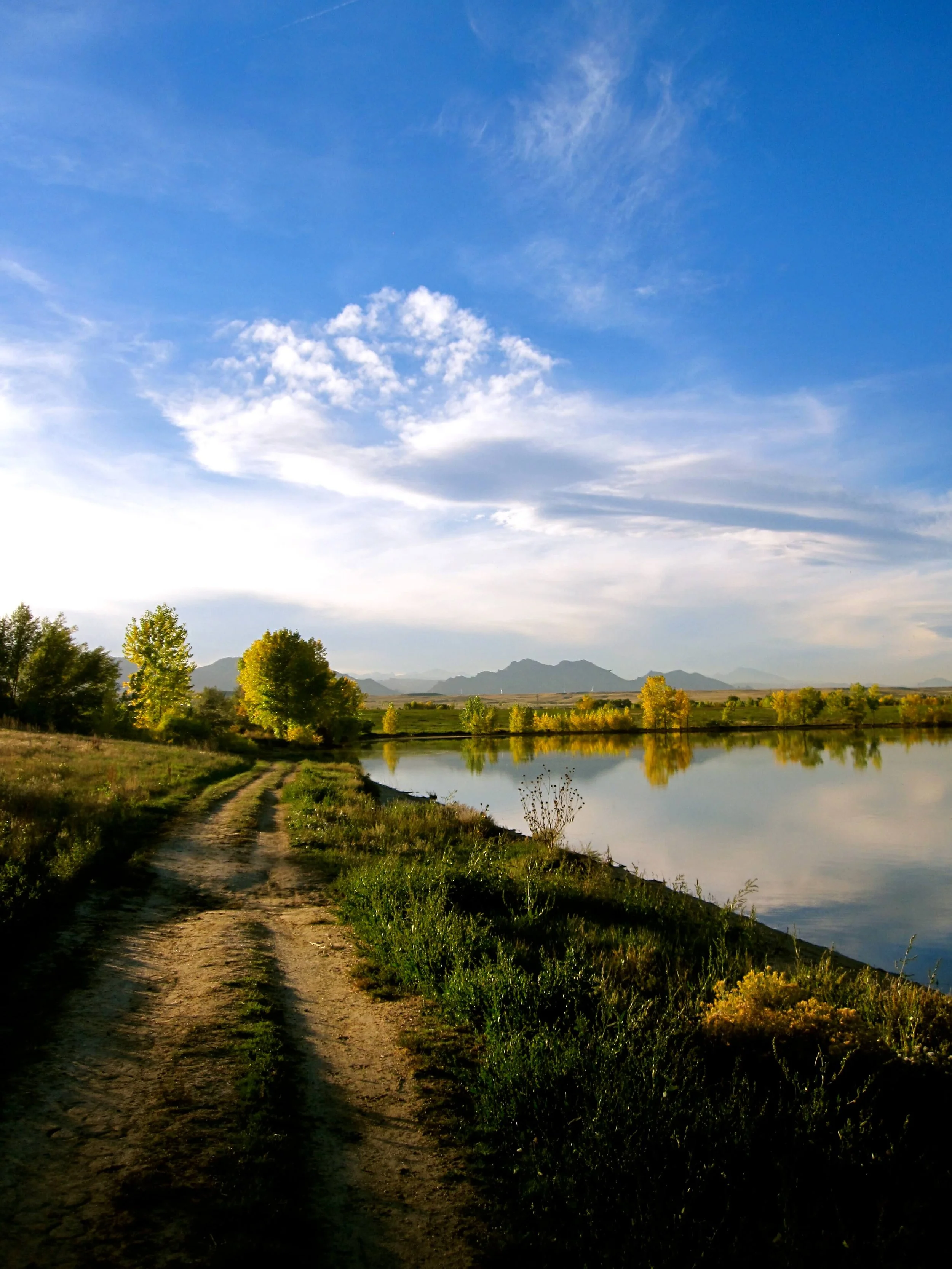Serene landscape with a dirt path along a calm river, surrounded by green trees, mountains in the distance, and a blue sky with scattered clouds.