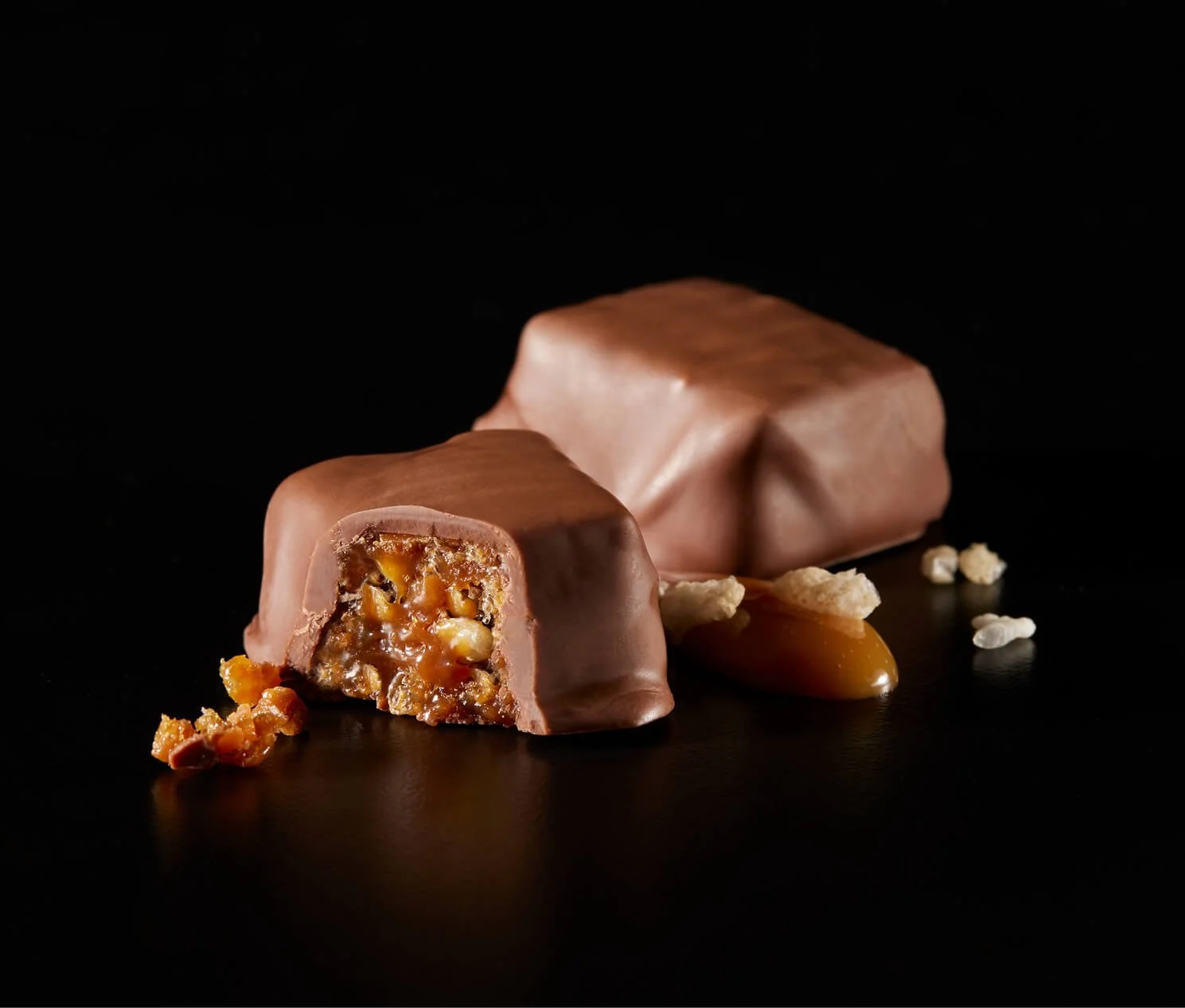 Chocolate-covered caramel confections with a nut filling, one cut open showing the inside and surrounded by caramel, nuts, and white nibs on a black background.