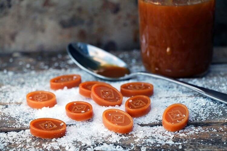 Caramel candies with a square design on top scattered on a rustic wooden surface with powdered sugar, a spoon with caramel sauce, and a jar of caramel in the background.