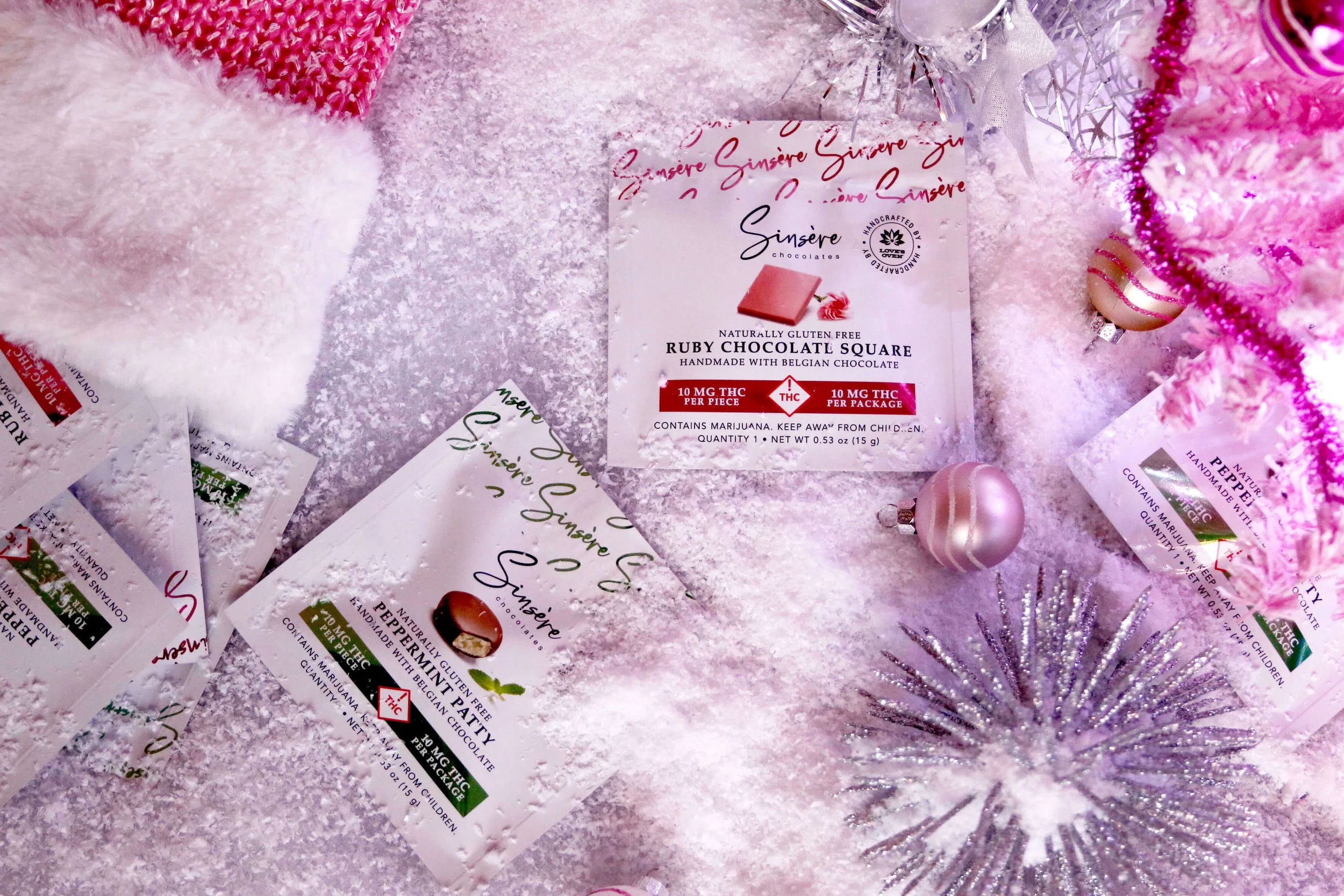 A display of pink and white Christmas ornaments, pink decorative trees, and fragrance tags in a snowy setting on a pink surface.