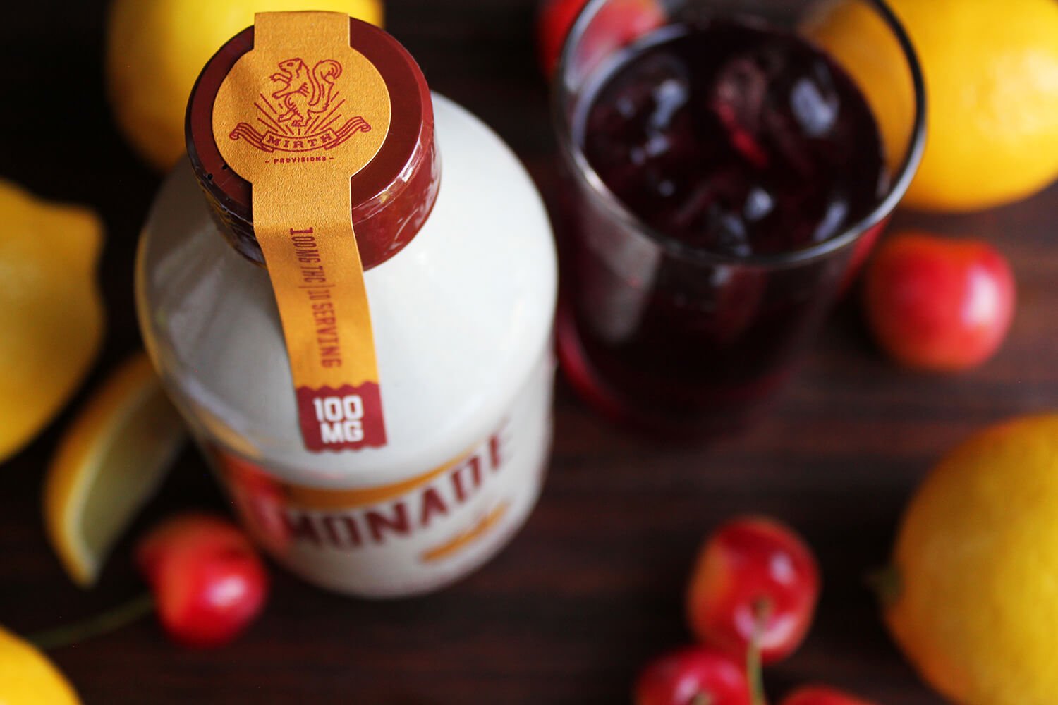 A bottle of cannabis CBD oil with orange and red label, a glass of cherry juice with ice, surrounded by lemons and cherries on a dark wooden surface.