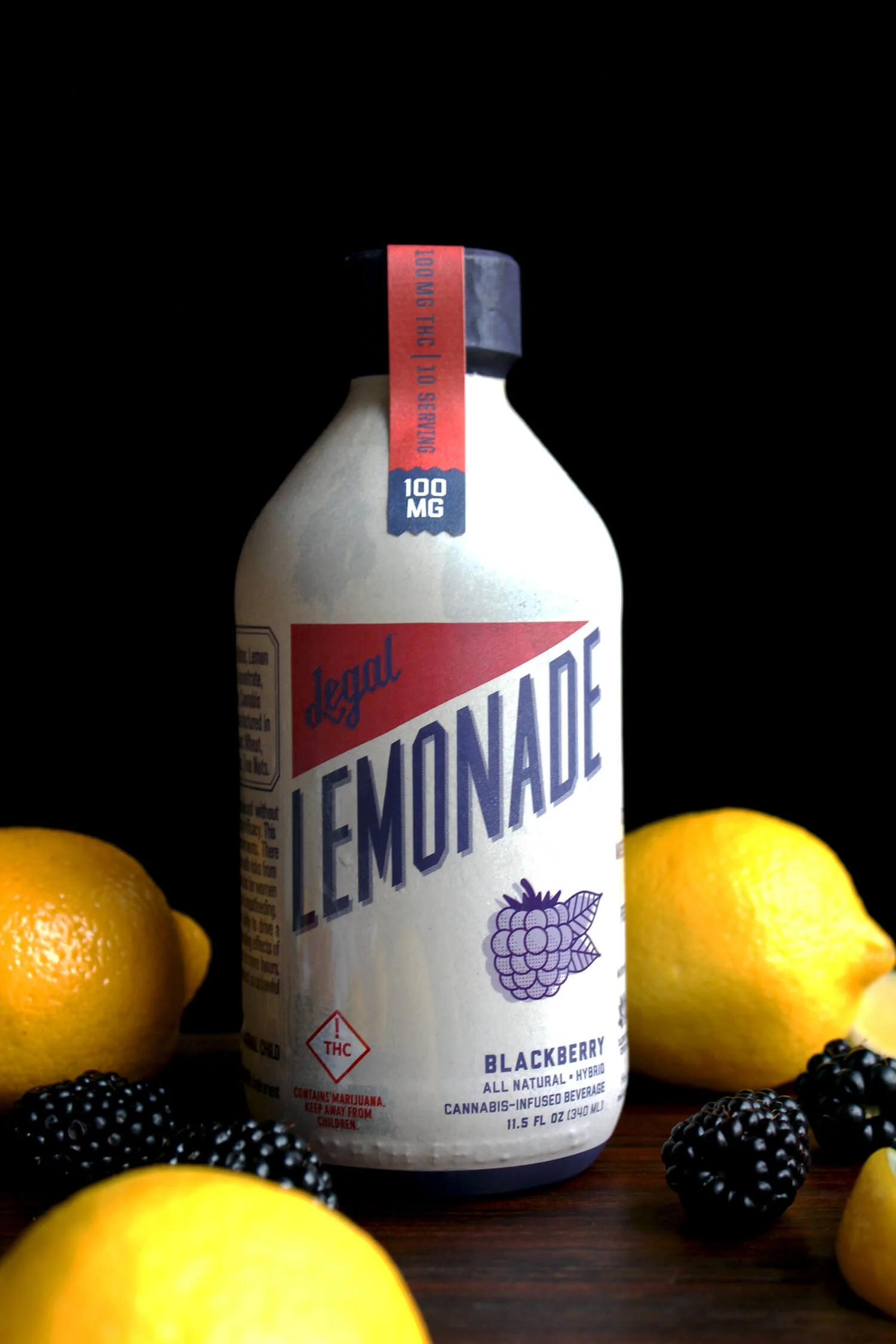 A bottle of blackberry cannabis-infused beverage called 'Lemonade' is centered on a wooden surface, surrounded by three yellow lemons and blackberries with some purple-colored berries, all against a black background.