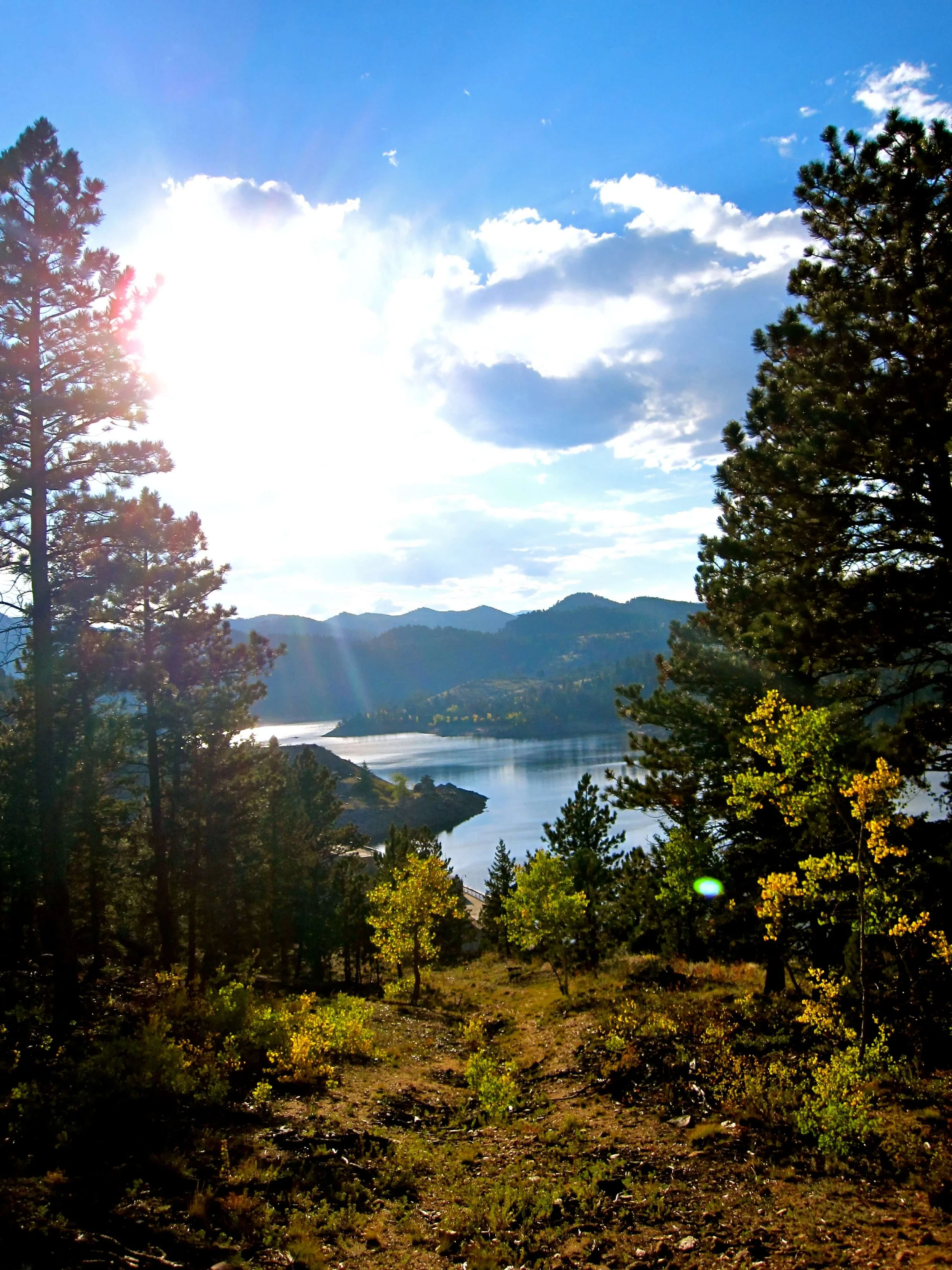 A scenic view of trees, mountains, a body of water, and a bright sun shining in a partly cloudy sky.