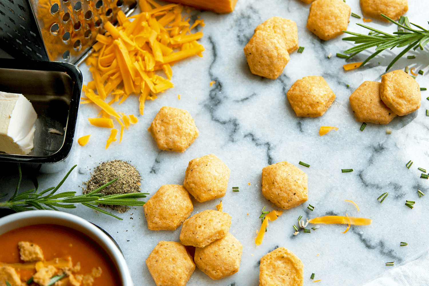 Uncooked chicken nuggets on a parchment paper, with shredded cheddar cheese, butter, herbs, and spices nearby, and a bowl of soup in the corner.