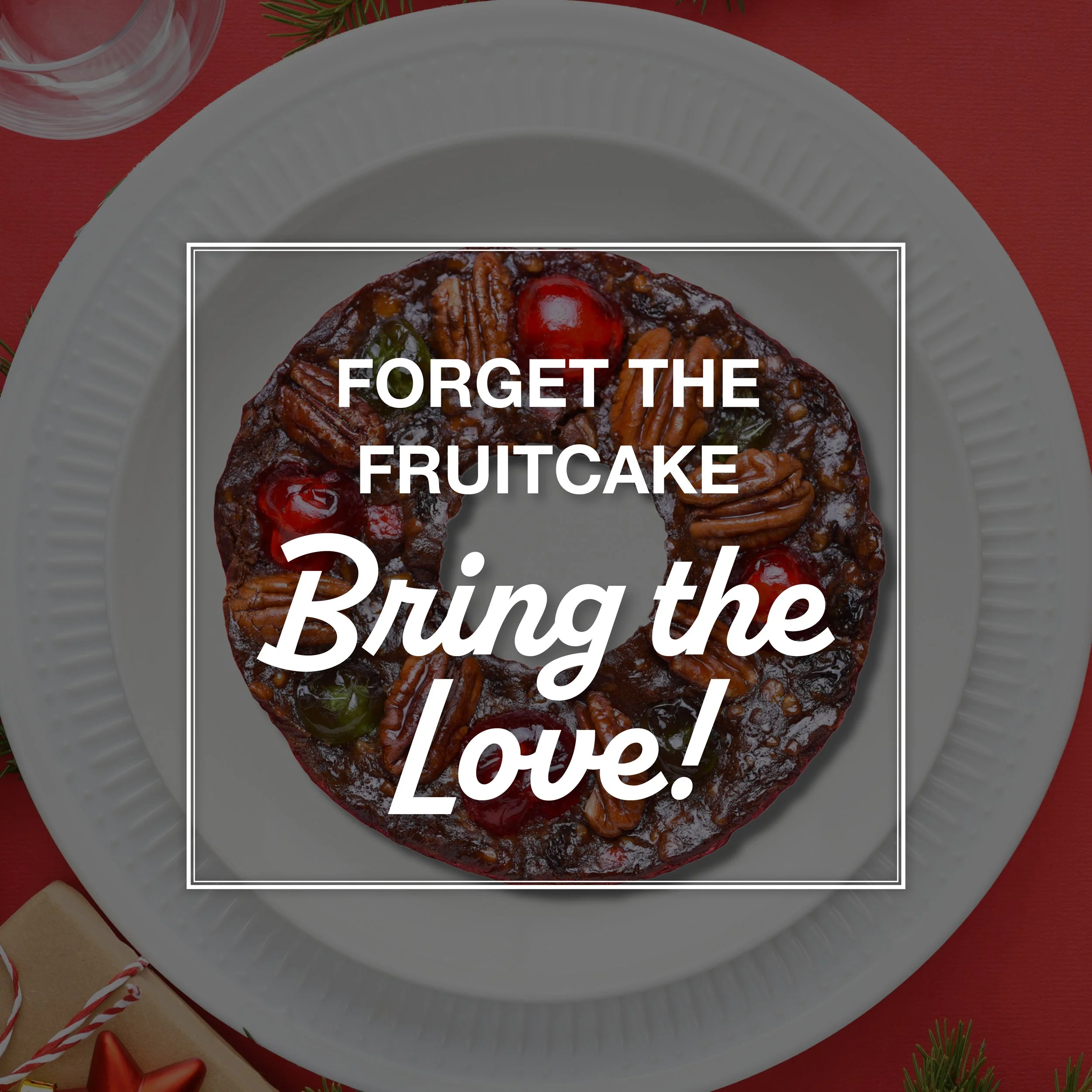 A fruitcake decorated with red cherries, pecans, and green candied cherries on a white plate. The background includes a red surface, holly sprigs, a glass of water, a wrapped gift with a red bow, and a star-shaped ornament.